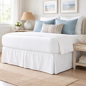 Può includere: Un letto con una balza bianca, biancheria da letto bianca, due cuscini azzurri e un cuscino rettangolare centrale con un motivo a righe beige e bianche. Il letto è posizionato tra due comodini in legno con lampade e decorazioni.