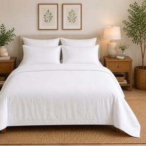 Puede incluir: Una cama con una funda nórdica blanca lisa y cuatro almohadas blancas a juego dispuestas en dos filas. La cama tiene un cabecero tapizado de color claro y está situada en el centro de una habitación con mesitas de noche de madera.