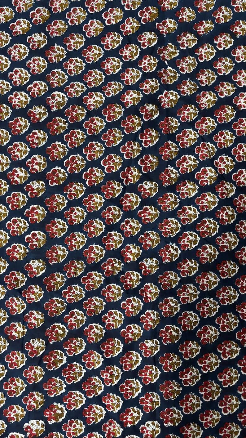 May include: Close-up of a fabric with a repeating floral pattern. The design features clusters of red and white flowers with green leaves on a dark blue background. The fabric is likely cotton or a similar material.