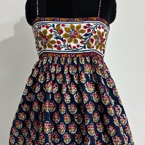 May include: A sleeveless dress with a floral print. The bodice features a white background with red, yellow, and green floral patterns. The skirt is navy blue with a repeating red and yellow floral design. The dress is displayed on a mannequin.