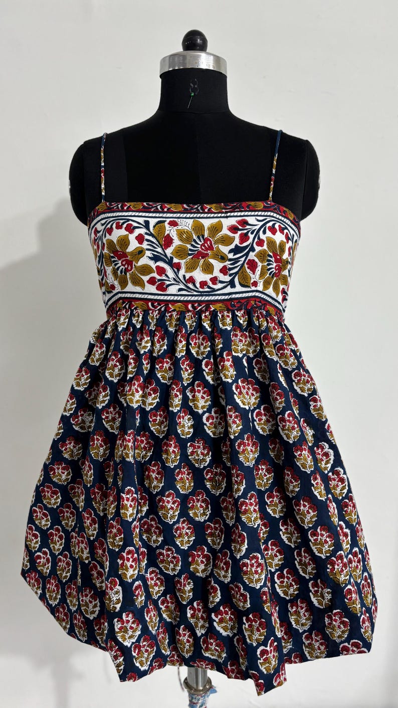 May include: A sleeveless dress with a floral print. The bodice features a white background with red, yellow, and green floral patterns. The skirt is navy blue with a repeating red and yellow floral design. The dress is displayed on a mannequin.