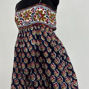 May include: A sleeveless dress on a black mannequin. The top features a white background with a floral pattern in red, yellow, and green. The skirt is navy blue with a repeating floral pattern in red and yellow.