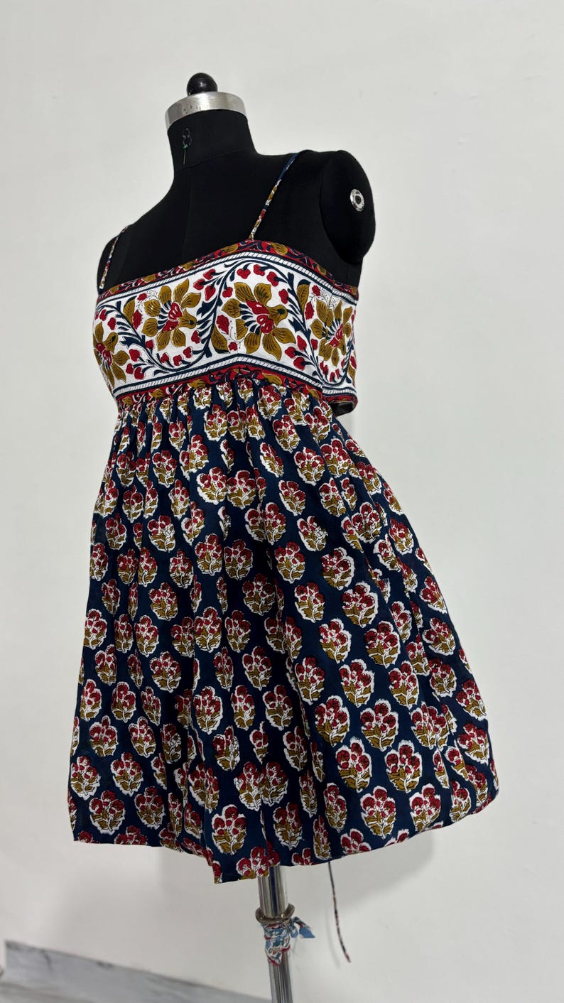 May include: A sleeveless dress on a black mannequin. The top features a white background with a floral pattern in red, yellow, and green. The skirt is navy blue with a repeating floral pattern in red and yellow.