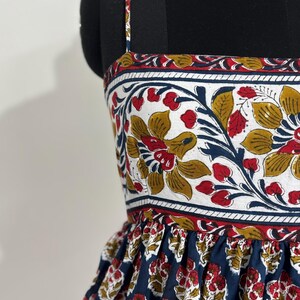May include: A sleeveless dress with a floral print. The bodice features a white background with red, yellow, and blue floral patterns. The skirt is navy blue with a repeating red and yellow floral design. The dress is displayed on a black mannequin.