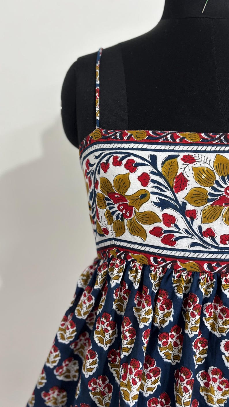 May include: A sleeveless dress with a floral print. The bodice features a white background with red, yellow, and blue floral patterns. The skirt is navy blue with a repeating red and yellow floral design. The dress is displayed on a black mannequin.