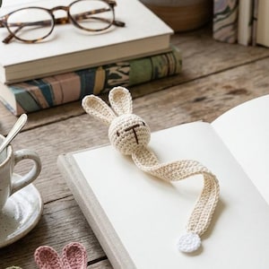 Könnte beinhalten: Ein Holztisch zeigt einen Häschen-Lesezeichen aus Häkelarbeit in einem offenen Buch. Zusätzliche Häschen-Lesezeichen aus Häkelarbeit in Grün und Rosa sind sichtbar. Eine Tasse Tee, eine Brille und Bücher befinden sich ebenfalls auf dem Tisch. Die Szene wird von natürlichem Licht beleuchtet.