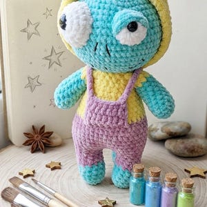 May include: A handmade crochet doll in a whimsical design. The doll is turquoise and yellow, wearing purple overalls. It has large white eyes and a yellow hood. Paintbrushes, star-shaped decorations, and small bottles of colorful glitter are arranged around the doll.