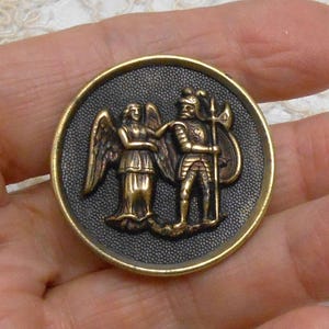 May include: A round, antique brass button with a detailed relief. The design features a winged angel and a knight in armor, holding a spear and shield. The background is a textured, dark gray, and the button has a gold-colored rim.