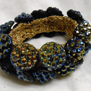 May include: A handmade bracelet featuring a gold crocheted center band, surrounded by black buttons and clusters of iridescent blue and gold beads. The bracelet has a unique, textured design.