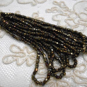 May include: A strand of small, metallic dark bronze beads with a faceted finish. The beads are strung together on a thin cord, creating a dense, textured cluster of reflective, dark-toned cylindrical shapes that catch the light.