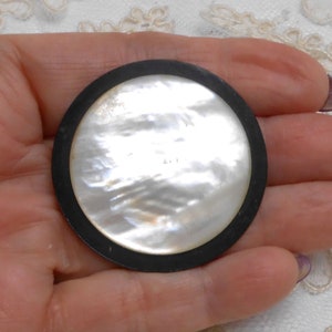May include: A round, white mother of pearl button with a black plastic rim.