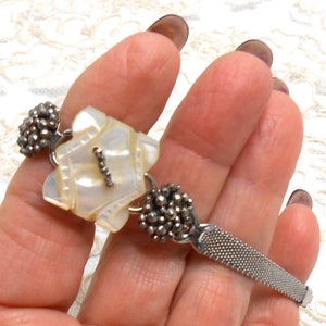 May include: A silver bracelet with a white mother-of-pearl charm and two silver bead clusters. The bracelet has a mesh band.