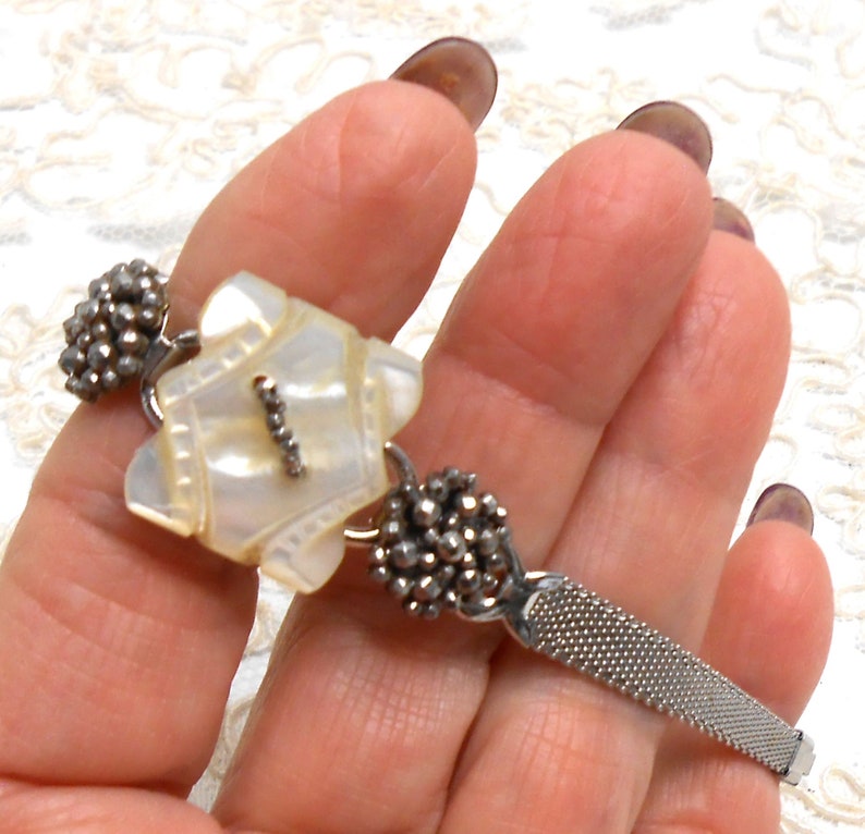 May include: A silver bracelet with a white mother-of-pearl charm and two silver bead clusters. The bracelet has a mesh band.
