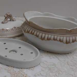 May include: A white ceramic soap dish set with a lid and a separate oval soap holder. The set features a decorative gold trim and a detailed handle on the lid. The soap dish is designed for bathroom use.