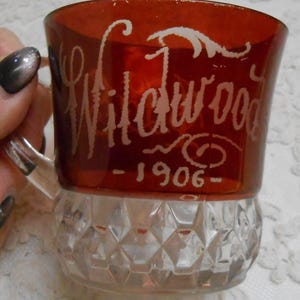 May include: A vintage red glass mug with a clear, diamond-patterned base. The mug features the etched word "Wildwood" and the year "1906" in white script. The handle is clear glass.