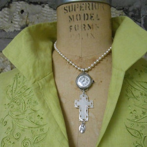 May include: A pearl necklace featuring a silver watch pendant and a carved cross pendant. The watch has a white face with black numerals and hands. The cross is white with an angel pendant. The necklace is displayed on a mannequin.