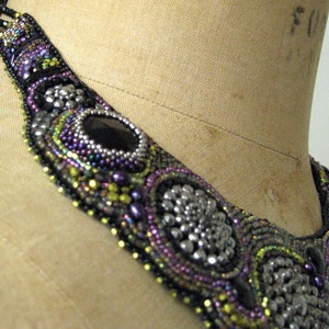 May include: An elaborate beaded necklace in black, silver, purple, and green. The design includes large, oval black stones and circular silver details. The necklace is displayed on a mannequin, showcasing the intricate beadwork.
