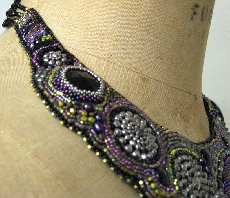 May include: An elaborate beaded necklace in black, silver, purple, and green. The design includes large, oval black stones and circular silver details. The necklace is displayed on a mannequin, showcasing the intricate beadwork.