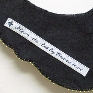 May include: A black fabric collar with a scalloped edge, decorated with small yellow and black beads. A white label with the text "Fleur-de-lis by Genevieve" is sewn onto the collar, featuring a fleur-de-lis symbol.