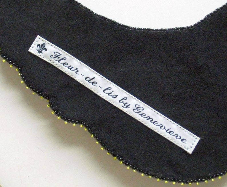 May include: A black fabric collar with a scalloped edge, decorated with small yellow and black beads. A white label with the text "Fleur-de-lis by Genevieve" is sewn onto the collar, featuring a fleur-de-lis symbol.