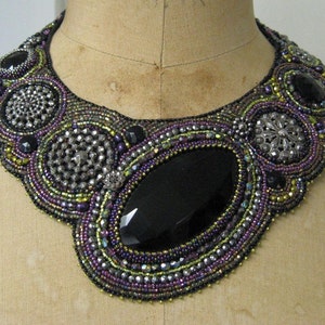 May include: A statement necklace, handmade with purple, black, silver, and gold beads. The bib-style design features a large, faceted black stone at its centre, encircled by detailed beadwork. The necklace is a striking piece of jewellery.