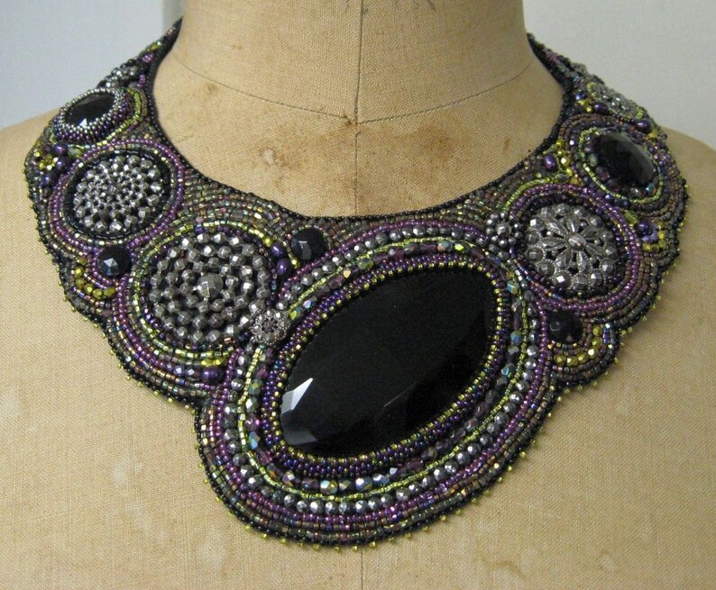May include: A statement necklace, handmade with purple, black, silver, and gold beads. The bib-style design features a large, faceted black stone at its centre, encircled by detailed beadwork. The necklace is a striking piece of jewellery.
