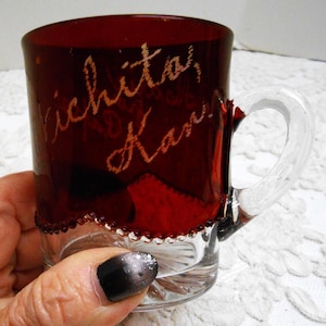 May include: A white ceramic mug with a black handle and the text "But First, Coffee" in a bold, black font. The mug is set against a blurred background, suggesting a cozy setting. The mug appears to be approximately 4 inches tall.