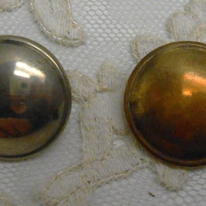 Könnte beinhalten: Zwei Metallknöpfe, einer silberfarben und einer goldfarben, liegen auf einer weißen Spitzefläche. Die Knöpfe sind rund und leicht gewölbt und reflektieren das Licht. Diese Knöpfe eignen sich zum Nähen und für Bastelprojekte.