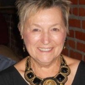 May include: A woman in a black top, adorned with a large, elaborate necklace. The necklace is made of black and gold beads, with a central medallion showing a figure. The woman has short, spiky hair and is smiling.