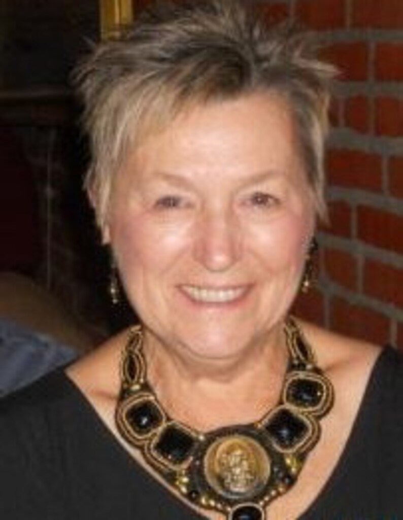 May include: A woman in a black top, adorned with a large, elaborate necklace. The necklace is made of black and gold beads, with a central medallion showing a figure. The woman has short, spiky hair and is smiling.