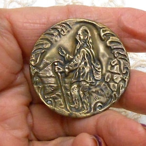 May include: A round, antique brass button featuring a detailed relief of a person with a staff, possibly a pilgrim, set against a landscape. The button has an ornate border and is held in a hand.