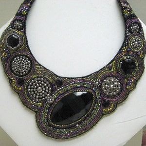 May include: A statement necklace with a large, black, faceted gemstone at its centre. The necklace is intricately beaded in black, purple, and iridescent green, with silver-toned details. The design is symmetrical and striking, with a bold, eye-catching aesthetic.