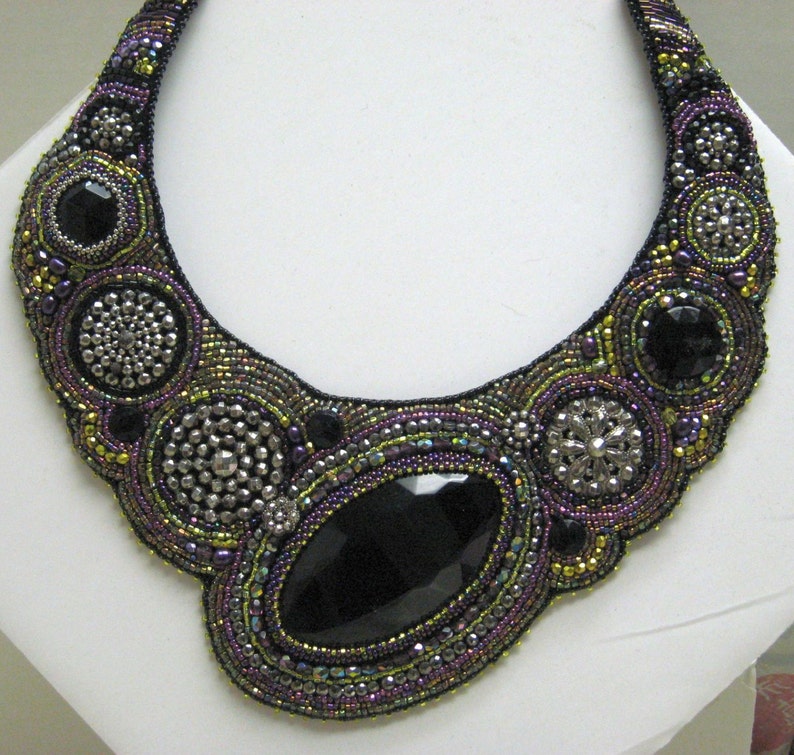 May include: A statement necklace with a large, black, faceted gemstone at its centre. The necklace is intricately beaded in black, purple, and iridescent green, with silver-toned details. The design is symmetrical and striking, with a bold, eye-catching aesthetic.
