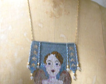 May include: A beaded necklace with a blue and gold patterned pendant featuring a portrait of a woman. The pendant is shaped like a rectangle with rounded corners and has a fringe of beads.
