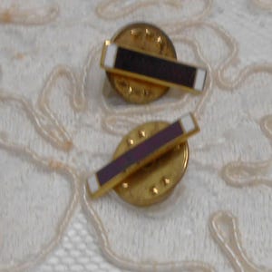 May include: Two gold-tone lapel pins, each featuring a horizontal rectangular bar with a dark purple centre and white enamel tips. The pins have circular gold-tone backings with a textured finish, designed to be secured with a standard pin clutch mechanism.