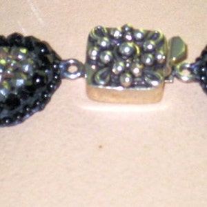 May include: Close-up of a black beaded necklace with a silver clasp. The necklace features two oval-shaped, black beaded sections with a central silver clasp. The beads are small and round, with a mix of black and green beads.