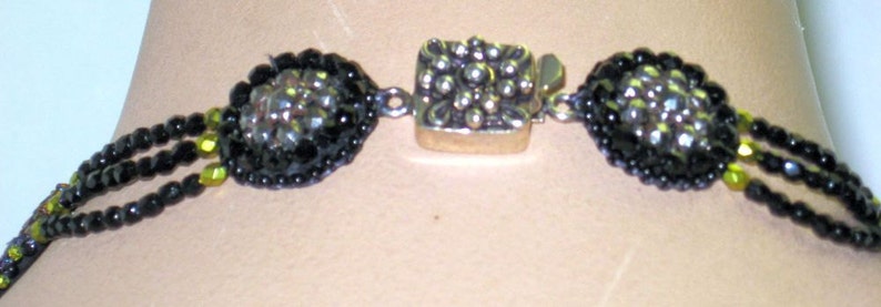 May include: Close-up of a black beaded necklace with a silver clasp. The necklace features two oval-shaped, black beaded sections with a central silver clasp. The beads are small and round, with a mix of black and green beads.