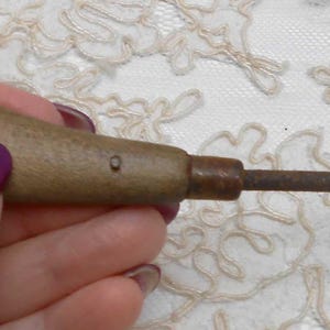 May include: A vintage rug hook tool with a wooden handle and a curved metal hook. The handle is a light brown color, and the metal hook has a dark, aged appearance. The tool is designed for pulling yarn through fabric to create rugs or other textile projects.