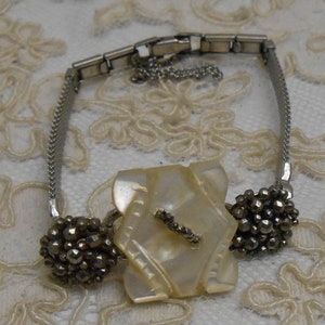 May include: A silver bracelet with a large, white, iridescent, geometric-shaped button and two smaller, round, silver buttons with a textured surface.