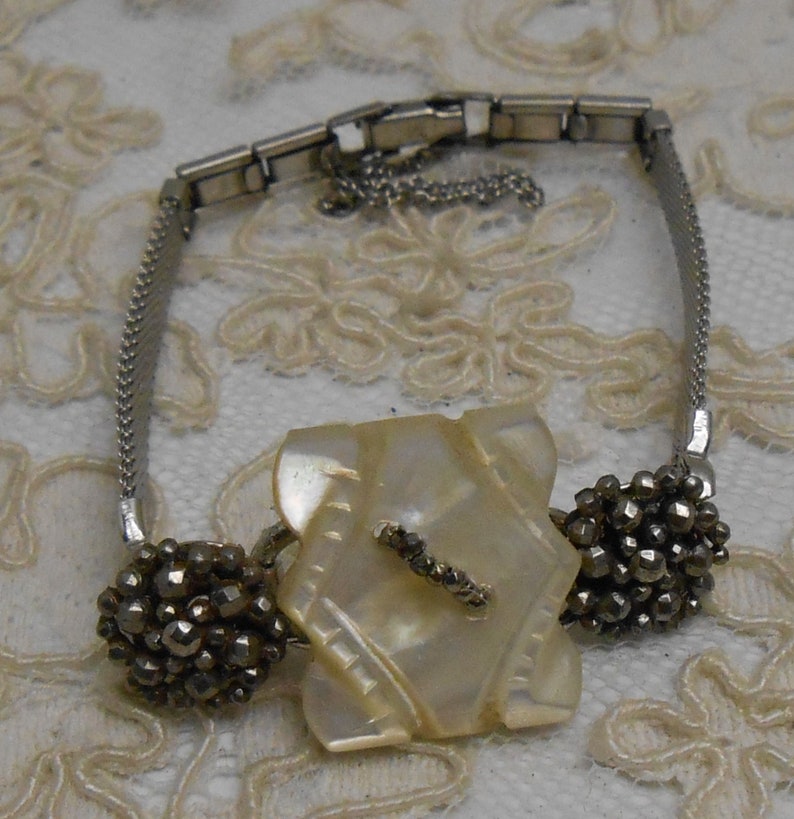May include: A silver bracelet with a large, white, iridescent, geometric-shaped button and two smaller, round, silver buttons with a textured surface.