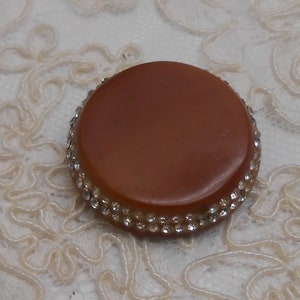 May include: A circular, brown button with a sparkling border of small, clear rhinestones. The button is set against a cream-coloured, lace-like fabric. The button's surface is smooth and has a slight sheen.