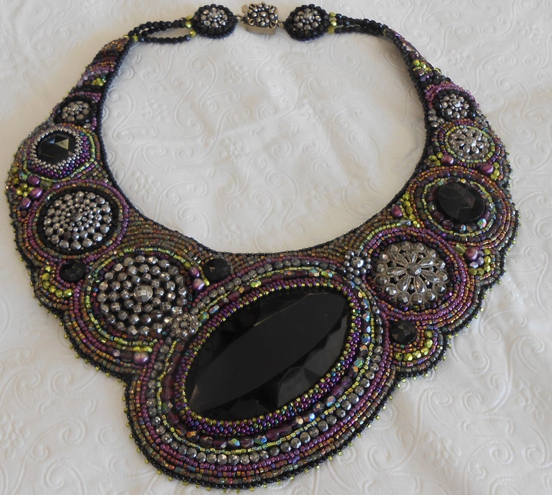 May include: An ornate beaded necklace with a large oval black gemstone at its centre. The necklace incorporates silver-toned details and iridescent beads in purple, green, and black. The clasp is silver-toned.