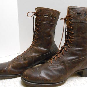 May include: A pair of dark brown leather lace-up boots featuring a rounded toe, a low heel, and a decorative cutout pattern along the upper shaft. The boots have thin brown laces and a weathered, textured finish on the leather surface.
