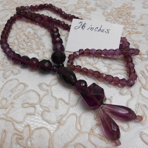 May include: A necklace featuring deep purple faceted beads of varying sizes and shapes, including round and elongated drops. A white tag with the handwritten text 26 inches is placed near the beads, indicating a total length of 26 inches.