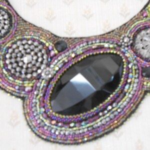 May include: A handcrafted beaded necklace with a bib design. The necklace features a large, faceted black gemstone at the centre, surrounded by intricate beadwork in shades of purple, silver, and iridescent colours. Additional silver embellishments add sparkle.