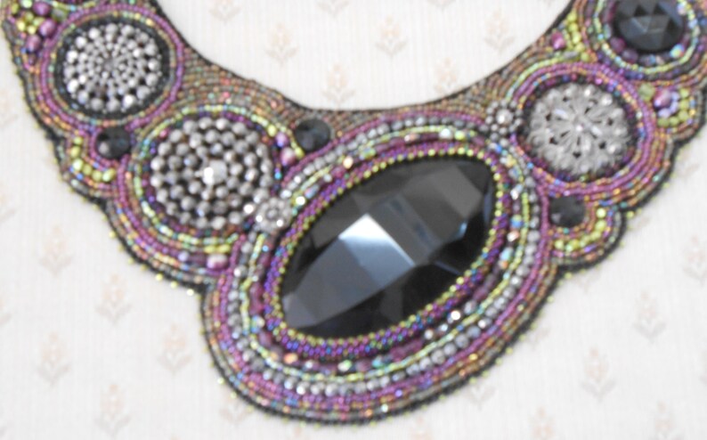 May include: A handcrafted beaded necklace with a bib design. The necklace features a large, faceted black gemstone at the centre, surrounded by intricate beadwork in shades of purple, silver, and iridescent colours. Additional silver embellishments add sparkle.