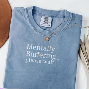 May include: A blue jean colored short-sleeve t-shirt with a crew neckline. The shirt features the white printed text Mentally Buffering... please wait. A gold-toned necklace with a small circular pendant rests on the collar of the shirt.