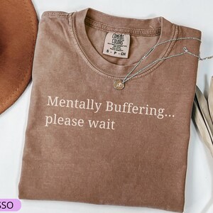 May include: Espresso-colored t-shirt with the text "Mentally Buffering... please wait" in white. A silver necklace with a pendant rests on the shirt. A brown hat and dried plant are also in the image.