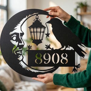 Pode incluir: Placa de endereço de metal preto com uma lua crescente com um rosto, uma lanterna e um corvo segurando uma estrela. A placa exibe o número 8908. O design é recortado do metal, criando uma silhueta única.