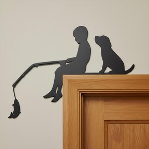 May include: Black metal silhouette of a child fishing with a dog. The child is seated, holding a fishing rod with a fish at the end. The dog sits attentively beside the child. The artwork is mounted on a wooden door frame.
